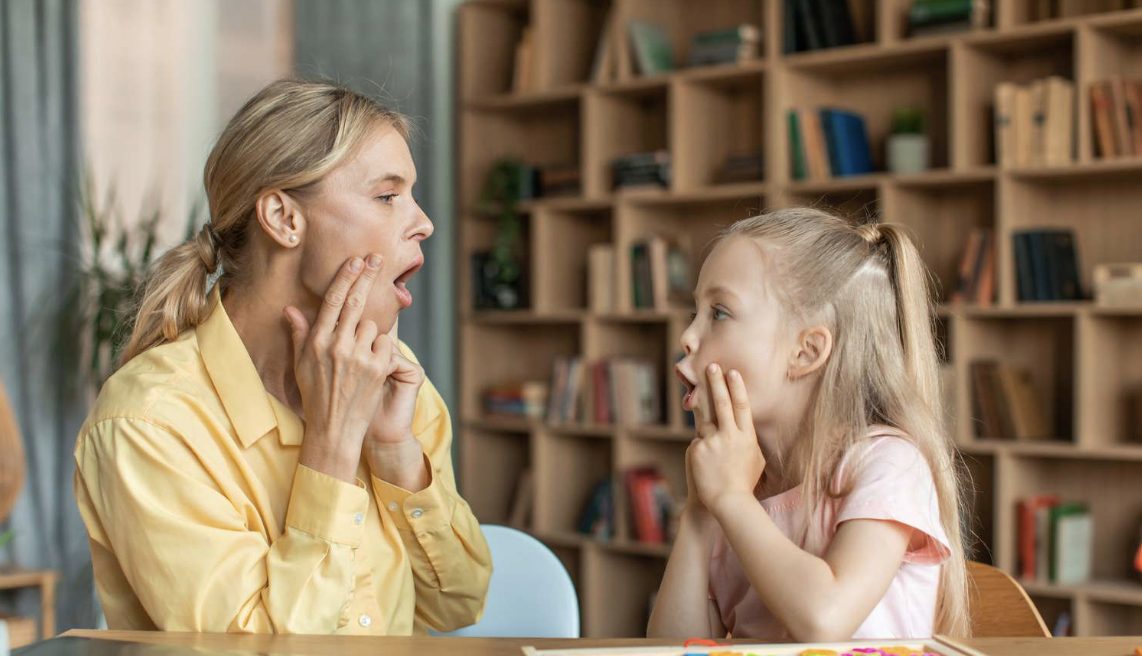 An adult and a young girl sit facing each other at a table, both touching their cheeks and forming open-mouth shapes.