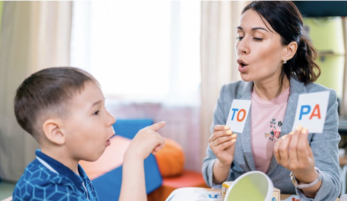 An adult holds two flashcards labeled 'TO' and 'PA' while a young boy points and opens his mouth.