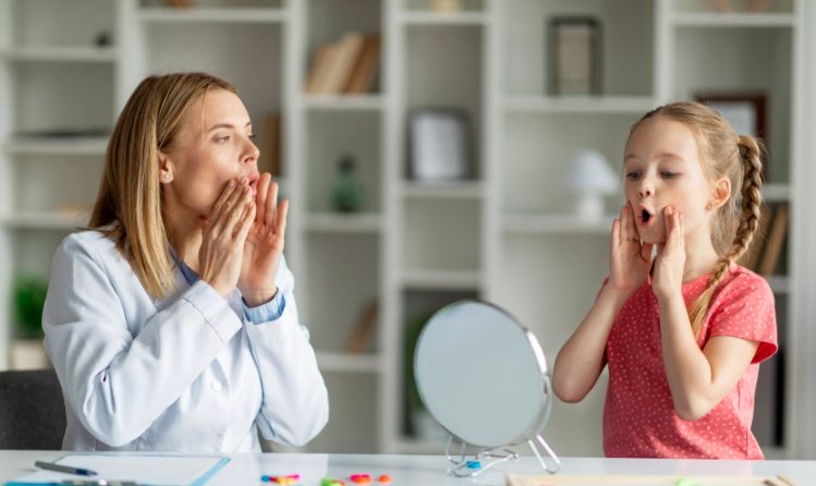 An adult in a white coat and a young girl sit at a table using a mirror while performing mouth exercises.